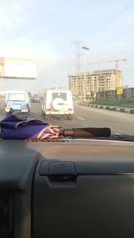 The video shows the interior of a car as it drives on a highway. In the foreground, the dashboard and steering wheel are visible, with a folded purple umbrella resting on the passenger side. The road ahead is populated with other vehicles, including a white SUV, a blue van, and several other cars and trucks. A large billboard for "CHIVAS REGAL" is prominently displayed on the left side of the road. In the background, there is construction activity with cranes and unfinished buildings, along with power lines and transmission towers. The weather appears to be clear and sunny. The camera perspective is from the driver's seat, looking forward.