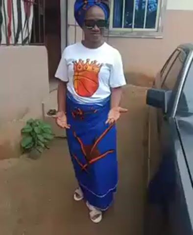 A medium shot shows a woman standing outside near a building and a dark gray or black car. The woman is smiling and facing the camera. She is wearing sunglasses, a blue head wrap, a white t-shirt with an image of a basketball with a crown on it, a blue wrap with orange designs around her waist, and white sandals. Her arms are slightly outstretched with her palms up. The building behind her is light beige and has bars on the window. A small green plant is on the ground to the left of the woman. The car is on the right, partially obscuring the background. The ground is a combination of dirt and concrete.