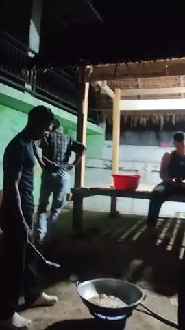 The video shows a group of people gathered outdoors at night. In the foreground, a person wearing a dark shirt and white slippers is standing near a portable gas stove with a wok on it. They are holding a spatula and appear to be cooking. Smoke is rising from the wok. Behind them, a man in a black shirt and jeans stands, looking towards the cooking area. Further back, two men are seated on a wooden bench under a thatched roof structure. One is wearing a dark tank top and the other a gray shirt. The environment appears to be a rural or semi-outdoor setting, possibly a yard or a communal cooking area, with dirt ground and a simple shelter. A bright light hangs above the seated individuals, illuminating the scene. Another person, whose face is obscured, is partially visible on the right side of the frame. The overall atmosphere suggests a casual, communal gathering, possibly for a meal.