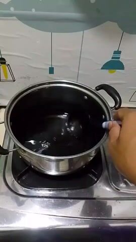 A hand is stirring a dark liquid in a pot with a ladle. The pot is on a stovetop, and the background is a wall with a decorative pattern.
