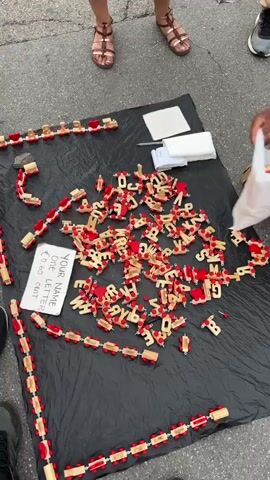The video depicts a street vendor kneeling beside a black mat spread on the ground. The mat is covered with small, wooden train cars, some assembled into trains and some separated into individual lettered pieces. A sign on the mat reads "YOUR NAME: 6 EURO LETTER: 60 CENT". The vendor is offering a train set with personalized letter cars spelling out the customer’s name. A person in a light green long-sleeved shirt takes some of the lettered train cars out of a plastic bag. A woman wearing a floral dress is standing in front of the display, holding a small child with brown hair and sandals on their feet. A man wearing white shorts and a light blue shirt holds a white plastic bag. The vendor is explaining the price of the product, stating that each letter costs 6 euros. The scene is set on a paved public area with other pedestrians walking in the background.