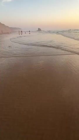 The video begins by showing a beach with soft waves gently rolling onto the shore during sunset. A few people and dogs can be seen walking along the waterline in the distance. As the video progresses, the camera focuses on the interplay of the waves and the wet sand, reflecting the warm hues of the setting sun. The sky displays a gradient of pastel colors, from soft pinks and oranges to a pale blue, with scattered clouds. A prominent rock formation stands out in the ocean to the left of the frame. The camera maintains a low angle, emphasizing the texture and movement of the water and sand.