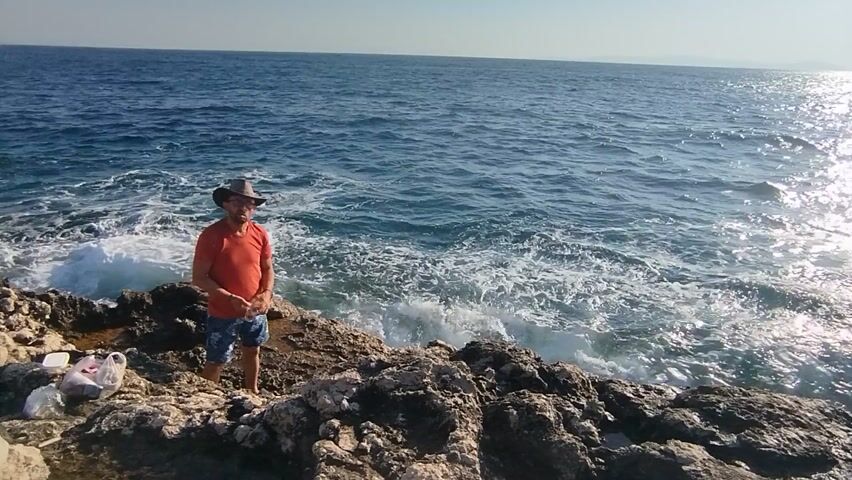 The video depicts an inverted perspective of a person standing on a rocky coastline, with the sea and horizon appearing above them. The person is wearing an orange t-shirt, blue patterned shorts, and a hat. They are positioned near the edge of the rocks, with waves crashing below them. The sun is bright, creating a strong reflection on the water's surface, suggesting it is midday. The setting is a rocky, natural landscape overlooking a vast expanse of blue ocean. The overall impression is one of a potentially adventurous or carefree moment, though the inverted view adds a surreal quality. There are no visible text elements in the video. The location context of Karaburun, Turkey, indicates a Mediterranean coastal environment.