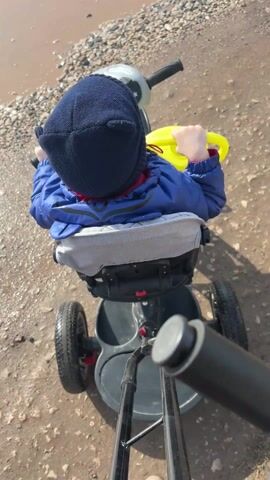 A video shows a toddler from behind, wearing a dark blue knit hat and a blue jacket with red trim, sitting in a black and gray tricycle-like device and holding onto a yellow object. The child is positioned at the edge of a large muddy puddle, bordered by light-colored pebbles and rocks. The tricycle has three wheels, a gray seat, and a push bar at the rear. A song is playing in the background.