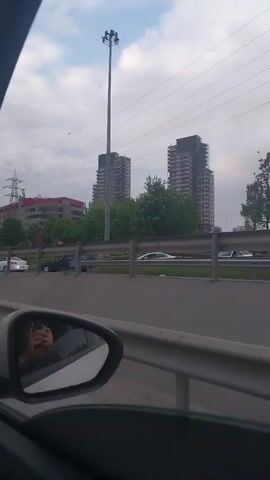 The video shows a view from inside a car driving on a highway. From the passenger side mirror, a person can be seen holding a phone, presumably recording. The road is busy with traffic in both directions. In the background, there are tall buildings, some under construction, and overhead power lines. The sky is overcast. The car eventually moves into traffic where other cars are visible in front. A unique shaped high-rise building is visible.