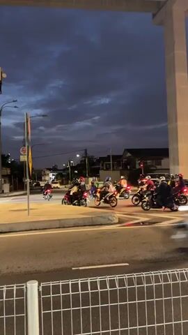 A group of motorcyclists ride through an intersection at dusk. The sky is dark with clouds, and streetlights and building lights are on. Several motorcycles are visible, some with riders wearing helmets. One motorcycle in the foreground turns right, while others are moving through the intersection in various directions. A large concrete overpass runs across the top of the frame. The road is paved, and there is a sidewalk with a yellow pole and signage. A metal fence is visible in the foreground.