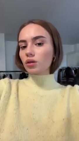 A young woman in a yellow sweater and black skirt appears to be filming a short video of herself. She is standing in a room that appears to be a dressing room, with clothing racks in the background. She looks directly at the camera. The camera angle changes, providing a brief view of a sofa with various items scattered on it. The camera returns to a closer shot of the woman's face.