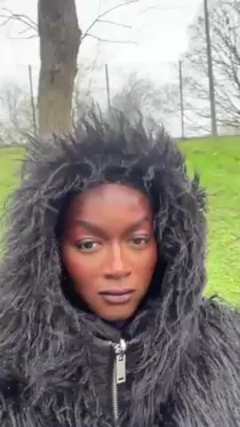 A person with dark skin and bold, stylized makeup is standing outside in an urban environment. The person is wearing a black hooded jacket with a shaggy faux fur trim around the hood. The person is looking directly at the camera with a neutral expression. The background includes trees, a grassy area, and a bridge or overpass structure. The sky is overcast and the lighting is muted, suggesting a cloudy day. The person appears to be standing still while recording a video.