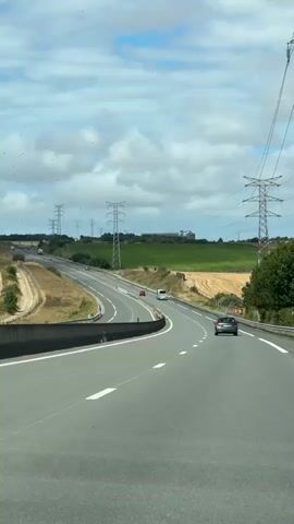 The video shows a car driving on a highway. The highway has multiple lanes and is bordered by grassy areas and fields. There are several electricity pylons visible along the side of the road, and a cloudy blue sky overhead. A few other cars can be seen on the highway, including one in front of the vehicle from which the video is being filmed. The terrain is somewhat hilly.