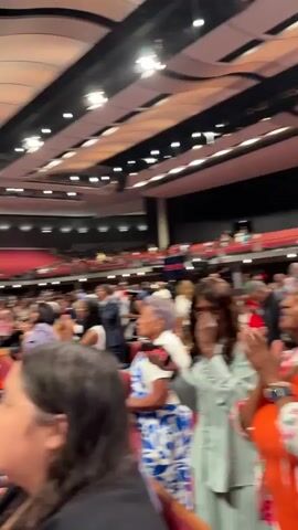 The video shows an indoor event, likely a church service, with a large audience seated in rows of wooden pews. The stage area features a large screen displaying an image of people standing in front of trees, and another screen displaying a man speaking. Lighting rigs and speakers are visible above the stage. The audience members, diverse in age and ethnicity, are mostly facing the stage. Some are standing, some are seated, and some appear to be praying or looking down. The overall atmosphere seems to be one of reverence or attention.
