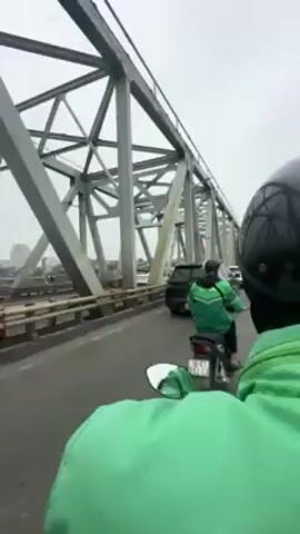 The video shows a view from the back of a motorcycle moving along a bridge. The bridge has a complex, gray metal truss structure overhead. Other vehicles, including cars and scooters, are also moving on the bridge. The sky is overcast. A couple on a white scooter passes on the left, both wearing helmets. The city skyline is visible in the distance.