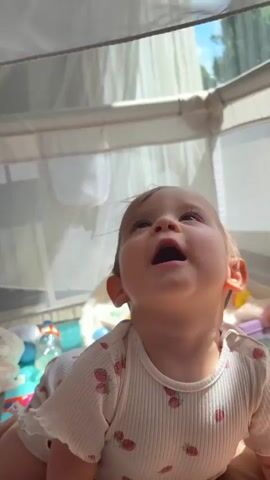 A baby is playing with a mosquito net in what appears to be a playpen. The baby is wearing a white onesie with red strawberry prints. The baby looks up and sticks out its tongue. The baby then grabs the mosquito net and pulls it down over its face, peeking out from underneath with a smile. The background is blurred but suggests an indoor environment with natural light coming from a window.
