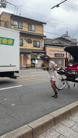 A person wearing a white short-sleeved top, white shorts, dark shoes, and a black bucket hat is seen from the right side of the frame, pulling a black rickshaw. The person is bent forward, gripping the two long shafts of the rickshaw and running forward to initiate movement. The rickshaw features two large, spoked wheels and a black canopy over a red seat. The scene is an urban street with buildings lining the background. A two-story yellow building with a fish decoration is visible first, followed by a multi-story grey building, and then a brown apartment building with external staircases. Overhead utility lines and poles are present. A black SUV is parked on the street at one point, and a white delivery truck passes by on the left. Towards the end of the clip, another person in a brown shirt walks into the frame from the left, briefly passing the rickshaw and its puller. The sky is overcast.