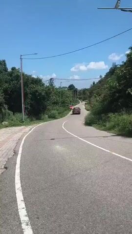 The video shows a winding paved road in a rural or semi-rural area. The road has a white line marking the edge. On the left side of the road, there is a sidewalk with some red and white striped poles. Lush green trees and vegetation line both sides of the road. In the distance, a red car is driving away from the camera. Overhead, power lines and streetlights are visible. The sky is blue with scattered white clouds.