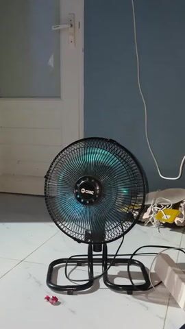 A black oscillating fan with a blue light on its central motor casing is positioned on a tiled floor in front of a wall. The fan's blades are spinning, creating a blur of blue light. The fan has a black metal stand with two circular feet. A white electrical cord extends from the back of the fan and runs across the floor. In the background, to the left, is a white door with a silver handle. To the right of the fan, on the floor, are various objects, including some white wires, a yellow item, and a small white box. The wall behind the fan is painted a muted blue color.