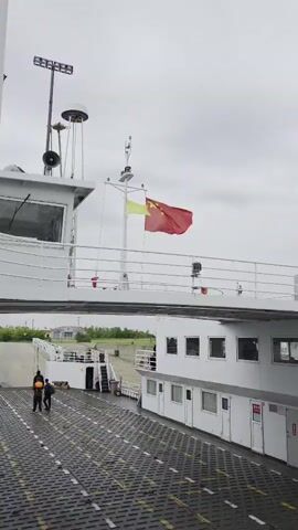 The video displays an exterior view of a large white boat or ferry under an overcast sky. A red flag with five yellow stars, the national flag of China, is prominently flown from a mast on the upper deck, with a smaller yellow flag directly beneath it. The boat's structure includes a bridge or command cabin with windows, railings, and various navigational equipment like a radar dome and antennas.

The main deck, covered with a textured, possibly non-slip, checkered surface, shows two individuals walking away from the camera at the start of the video. Another person is visible standing near the railing on a lower section of the boat. In the background, what appears to be land or a dock with some green vegetation and structures can be seen, indicating the boat is either docked or in close proximity to the shore. The scene is set during the daytime, though the sky is cloudy. The flags are gently waving, suggesting a light breeze. No discernible text is visible on the boat itself.