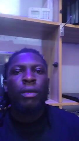 The video shows a medium shot of a Black man with dreadlocks and a beard, wearing a black shirt. He's looking directly at the camera with a neutral expression. Behind him is a shelf with various items. There is a Lenovo box and an electronic device with several buttons visible. The lighting appears to be somewhat dim and has a bluish tint. The man is the primary focus of the video, and he does not appear to be engaged in any particular action other than holding the camera. The video has no sound.