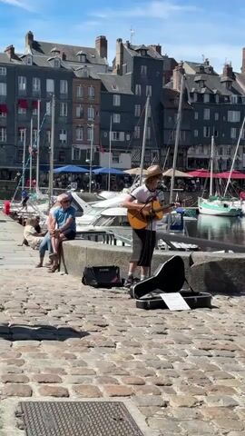 The video shows a street musician playing an acoustic guitar while standing on a cobblestone path. A guitar case lies open on the ground in front of him, with sheet music inside. In the background, a harbor is visible with boats docked and buildings lining the waterfront. Two people are sitting on a stone wall to the left of the musician, appearing to listen. Another woman walks by on the right. The weather is sunny.