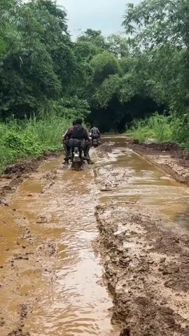 Two men ride a motorcycle down a heavily muddy and rutted dirt road, surrounded by lush green foliage. The first man is driving, while the second man is riding as a passenger. The road is completely covered in deep, brown mud and water. The trees and plants on either side of the road are vibrant green, indicating a tropical or subtropical environment. The sky is overcast. The video also shows a close-up of the driver's hands on the handlebars, with a water bottle tied to the side of the motorcycle.