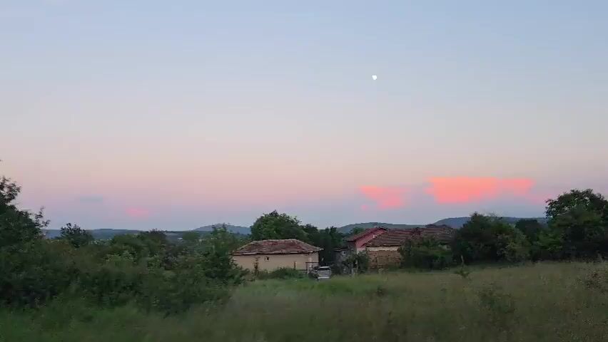 This is a video clip of a rural landscape at dusk. The scene features rolling hills covered in vegetation, with a few houses visible in the middle ground. The sky is a soft gradient of pink and pale blue, with a full moon or a bright planet visible as a small white orb. The time of day is clearly evening, as indicated by the soft lighting and the presence of the moon. The weather appears calm and clear. The location cues suggest a natural, undeveloped area, possibly a village or a small town in the countryside. There are no people or specific objects of interest, other than the houses and the natural elements. The overall mood is serene and peaceful, with the twilight colors contributing to a sense of tranquility. The video appears to be shot from a moving vehicle, as evidenced by the slight blur and the way the landscape shifts. No text is visible in the video.
