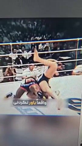 Two men are engaged in a wrestling match within a ring. One man, wearing black shorts with red trim and blue-accented gloves, is on top of the other, who is wearing black shorts with red and green accents. The man on top has the other man in a hold, lifting him and appearing to slam him down. The action is captured in a series of still frames that create a sense of dynamic movement and impact. The background suggests a crowd or spectators watching the event. The scene is intense and suggests a combat sport scenario, likely mixed martial arts or professional wrestling, given the nature of the hold and the apparent force involved.