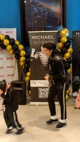 Two male individuals, one appearing to be an adult and the other a child, dressed in black sequined jackets and pants reminiscent of Michael Jackson's attire, are engaged in a dance performance. They are positioned in front of a banner that reads "MICHAEL JACKSON" and features an illustration of the singer. A balloon arch made of black and gold balloons frames the left side of the scene. The background includes a blue wall and a movie poster. The adult is gesturing towards the child, who is performing a moonwalk.