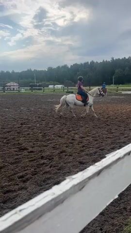 The video shows a horse riding arena with a rider on a white horse. The ground is covered in dirt. There is a white fence in the foreground. In the background, there are trees and a cloudy sky. The rider is wearing a helmet and orange riding pants. The horse is moving at a trot. Another person on a horse is visible in the distance.