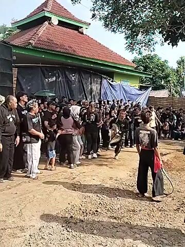 The video shows a traditional performance in what appears to be a rural Indonesian setting. A crowd of people is gathered outdoors in a dirt-covered area, with buildings, trees, and other foliage visible in the background. The focal point is a man dressed in traditional clothing, including a head wrap and decorative pants. He appears to be dancing or performing a ritual, as he moves around holding something. Other people are watching the performance, some recording with their phones, and others standing around. The scene is filled with activity and seems to be a communal celebration.