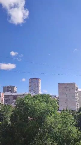 The video displays a view of a city skyline on a clear day. The sky is a bright blue with scattered white clouds. In the foreground, lush green trees obscure the lower portion of the frame. Behind the trees, several tall buildings are visible. On the left, a cluster of buildings, likely apartment blocks, are present. To the right of these, a taller, modern-looking building with a distinct facade stands out. Further to the right, another, wider apartment building with visible windows is seen. The top of a construction crane can be seen on top of the central tall building, indicating ongoing construction. Thin, dark lines, possibly power lines or wires, stretch horizontally across the sky in the middle ground. The overall impression is an urban landscape under a sunny sky.