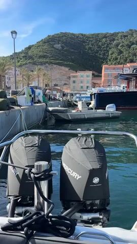 The view from a boat in a harbor pans right to show a dark blue yacht called “Sloopy.” The shot is from behind the rear rail and two covered Mercury outboard motors. Other boats, mostly smaller, are moored along a concrete pier. Buildings are visible on shore, with a backdrop of a tree-covered hill under a blue sky. A plane flies overhead.
