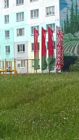 The video shows a view of a multi-story apartment building. In the foreground, there is a grassy area with some wildflowers. The building is painted in pastel colors, with alternating sections of light blue, grey, and pale yellow. There are many windows on each floor, some with balconies. To the right of the building, a portion of a billboard is visible. In the background, there is a clear blue sky with some scattered clouds. The camera appears to be moving, as the perspective shifts slightly throughout the short clip. The overall scene depicts a residential area in what appears to be a town or city.