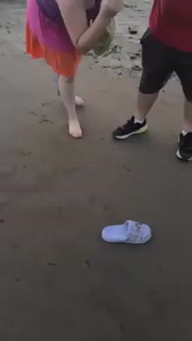 The video shows a woman on a sandy beach, near a man wearing sneakers and a red shirt. The woman is wearing a purple, pink, and orange dress and appears to be putting on a light blue sandal that had been lying on the ground. The beach is sandy with scattered debris.