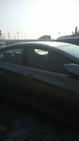 The camera pans across the front and side of a gray Hyundai Elantra parked in a lot. A few other cars are parked nearby, including an SUV. Buildings can be seen in the background on the left. The sky is hazy.
