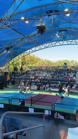The video shows a pickleball match underway on an outdoor court. There are four players on the court, two on each side of the net. They are all wearing athletic attire and holding pickleball paddles. A referee is standing to the side of the court observing the game. Spectators are seated in the bleachers, watching the match. The court is under a blue metal canopy that provides shade. The sky is clear and blue. Banners are strung across the back of the bleachers. Advertising for brands such as Joola and Franklin can be seen on the court and in the spectator area. The camera is positioned at a low angle, looking up at the court and spectators.