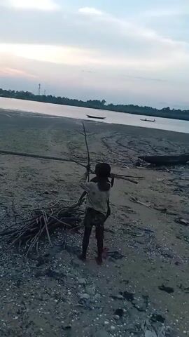 The video shows a coastal scene at dusk. Three young children, dark-skinned and scantily clad, play on a sandy beach. Two of them are holding sticks. There are piles of driftwood scattered on the beach. In the background, the sea is calm and there are several small boats visible. The sky is overcast.