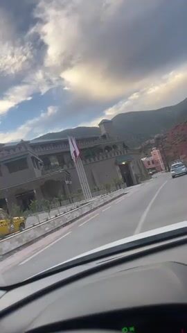 From the perspective of a car driving down a paved road, the scene captures a building with a Moroccan flag waving in the breeze. The building is tan-colored with a green trim on the roof. Behind it, hills rise against a partly cloudy sky. A couple of cars are driving in the opposite direction. There is a low stone wall between the road and the building. Other structures, including one that is pink, dot the landscape on the side of the road. The vehicle moves forward along the road, and the scene evolves to show more of the red dirt hills.