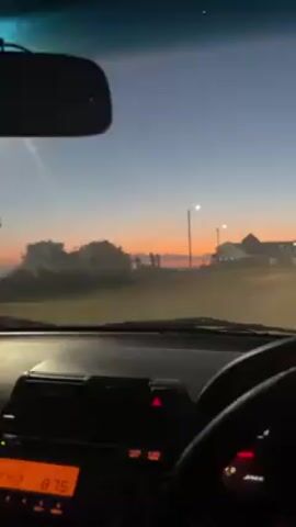 A view from inside a car driving down a road at dusk. Buildings are visible in the distance, silhouetted against the orange and blue sky. The dashboard, steering wheel, and speedometer of the car are visible. The speed of the car is indicated as 35 kilometers per hour. The radio is turned on.