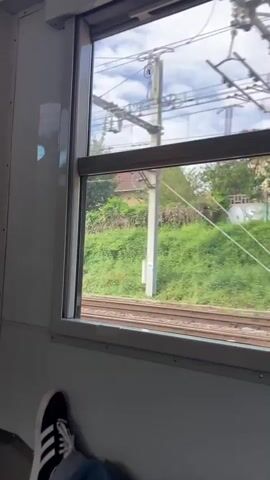 The video shows a view from the window of a moving train. The scene outside the window consists of a railway track, a grassy embankment, and various buildings and foliage in the background. The sky is visible with patchy clouds. Inside the train, the interior walls are painted white, and a person's leg wearing an Adidas sneaker is visible resting near the window.