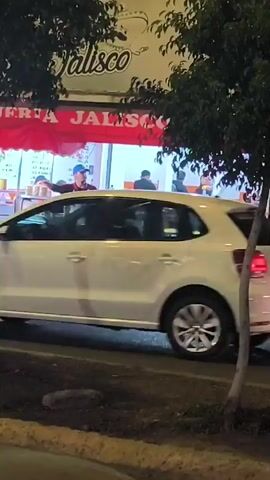 The video shows a street scene at night with a restaurant named "Torteria Jalisco" visible. People are sitting at tables inside the restaurant. A man wearing an apron and a baseball cap stands near a food cart outside the restaurant. Cars pass by on the street. There are trees framing the view on both sides. The setting appears to be an urban area.