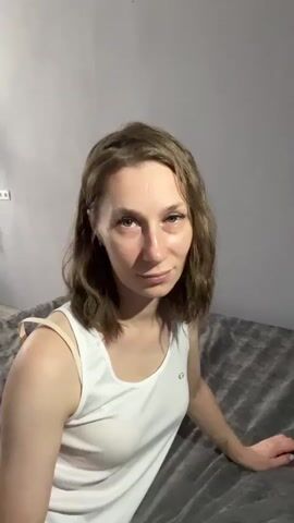 A young woman with shoulder-length brown hair and a white tank top is sitting on a bed. She looks into the camera and then brings her hands together to form a heart shape over her face. The background is a plain wall and a grey bedspread.