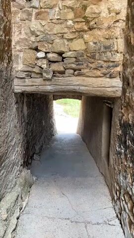 A stone passageway with a rough ceiling of wooden beams is shown from a fixed perspective. The passageway appears to be outdoors or open to the elements at the end. The walls are constructed of rough stones and mud or plaster. The floor is paved with concrete. The camera is static and the video is continuous with no discernible action.