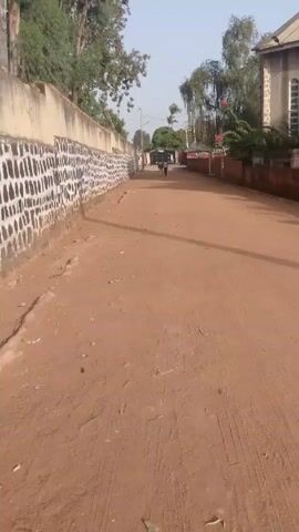 A video depicts a dirt road with two people walking away from the camera. The road is lined on either side by walls and buildings, with trees and foliage visible in the background. The people are walking towards a large gate or entrance. The sky is overcast.