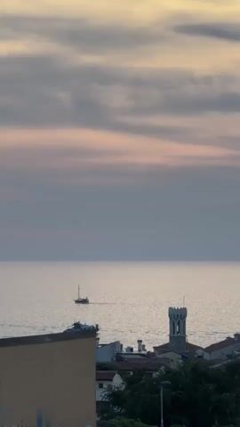 A vertical, eye-level, medium shot shows a seascape at dusk with a sailboat in the distance. The sky is a muted lavender and pink, with streaks of gray clouds. The sea is a calm, silvery expanse, with faint ripples. A small, dark sailboat with a single mast sails horizontally across the frame from left to right. In the foreground, the top of a weathered, mustard-yellow building occupies the lower left corner of the frame, with graffiti visible on its side. Beyond the building, a cluster of buildings with red-tiled roofs is visible, dominated by a tall, square tower with battlements. Trees and a street lamp are also visible in the mid-ground. The overall mood is tranquil and serene.