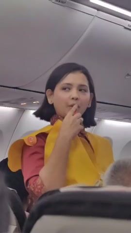The video shows the interior of an airplane cabin during what appears to be a safety demonstration. A flight attendant, wearing a yellow life vest over her uniform and a patterned scarf, stands in the aisle and gestures with her right hand. She has short, dark hair and is looking to her left. Behind her, an exit sign with arrows is visible on the ceiling. Other passengers are seated in the foreground, with their heads and shoulders visible. Another flight attendant in a similar yellow vest is visible further down the aisle. The lighting is bright and typical of an aircraft cabin.