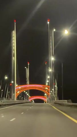 A first-person perspective video captured at night shows a vehicle driving over a bridge. The bridge is illuminated by orange and white lights along its structure, with two distinct archways visible in the distance. Tall, slender poles with red lights at their tips stand on either side of the roadway, supporting cables that ascend into the dark sky. Streetlights with a warm glow also line the bridge. The road surface is asphalt with visible lane markings. The overall scene conveys a sense of movement and the illuminated architecture of the bridge at night.