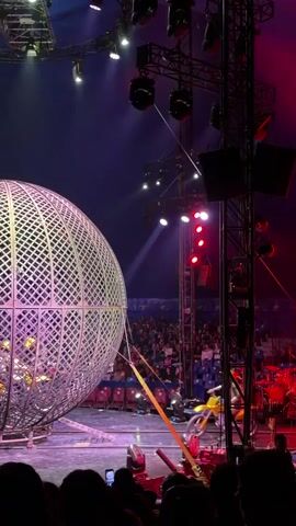 A motorcyclist in yellow gear prepares to enter a large metal sphere. The sphere has a diamond pattern lattice and is on a circular track. Inside the sphere, several other motorcycles are visible. The scene takes place in a dimly lit arena with a dark blue ceiling adorned with spotlights and rigging. A crowd is visible in the background, seated in stadium-style seating. The motorcyclist is positioned to the right of the sphere, with the motorcycle facing the entrance. The camera is positioned at eye level, capturing the event from the perspective of an audience member.