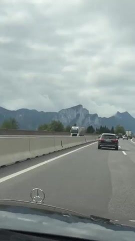 The video shows a view from inside a car on a highway, with a mountain range in the background. The sky is cloudy. On the highway, there are several cars, including a white truck and a dark SUV. The car from which the video is filmed appears to be a Mercedes-Benz, indicated by the emblem visible on the dashboard. The car is moving forward on the highway.