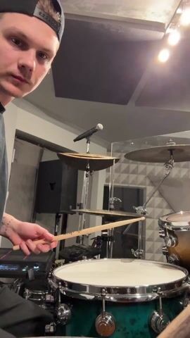 The video shows a young man with light skin playing drums. He is wearing a black baseball cap and a black shirt. He is smiling and sticks his tongue out at the camera. He has tattoos on his left forearm. He is playing a drum set with a green snare drum and a brass-colored tom. There are several cymbals visible. A microphone stands in front of the drums. The background consists of a room with gray walls, soundproofing panels, and studio equipment. The lighting is bright, with several lights visible in the ceiling. He plays quickly and energetically, hitting the snare drum and cymbals.
