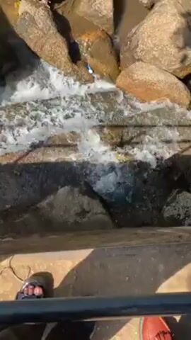 From an elevated perspective, the video shows a small, rocky stream. The water flows rapidly over and between large, irregularly shaped rocks. Some of the rocks appear smooth and worn, while others are rough. There are hints of vegetation in the background. The edges of the stream are lined with concrete and metal railings are visible. The bottom of a person's feet are visible, they're wearing sandals.