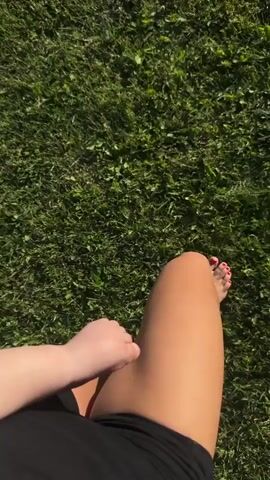 A first-person perspective video shows a person walking barefoot on green grass. The person is wearing black shorts. The sun is shining and casting shadows. The camera is focused on the person's legs and feet.